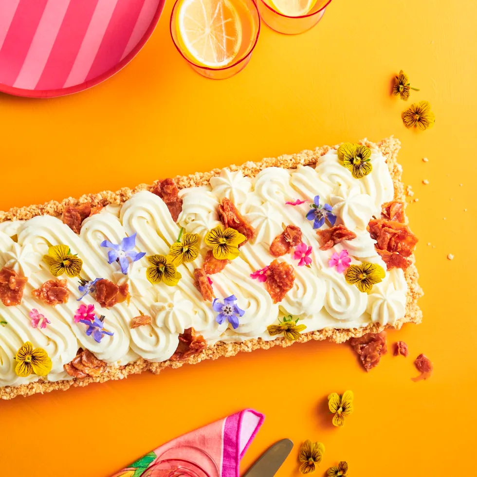 A rectangular cheesecake with a swirled pattern cream top with scattered edible flowers to decorate. On a yellow background