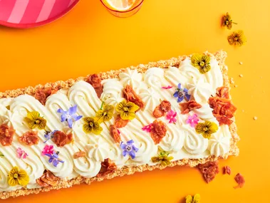 A rectangular cheesecake with a swirled pattern cream top with scattered edible flowers to decorate. On a yellow background