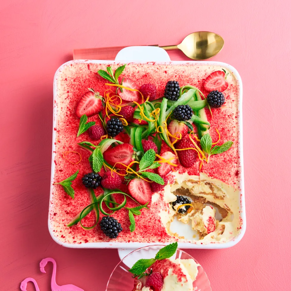 Tiramisu with a dusting of strawberries, with ribbons of cucumber and sprigs of mint on top in a square white baking dish sitting on a pink background