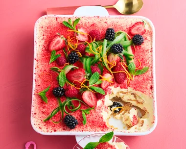 Tiramisu with a dusting of strawberries, with ribbons of cucumber and sprigs of mint on top in a square white baking dish sitting on a pink background