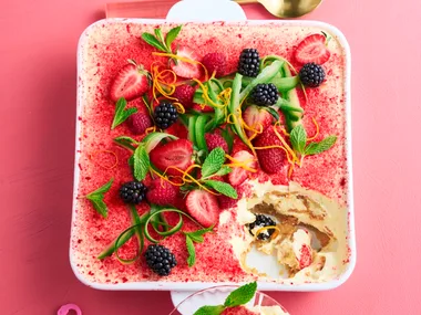 Tiramisu with a dusting of strawberries, with ribbons of cucumber and sprigs of mint on top in a square white baking dish sitting on a pink background