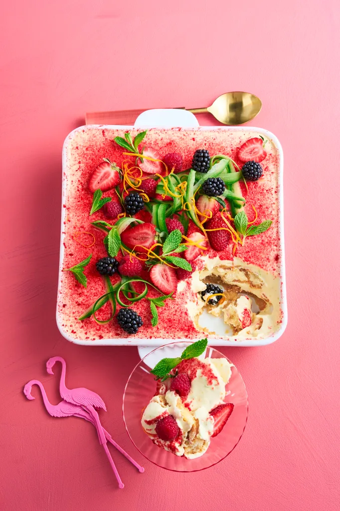 Tiramisu with a dusting of strawberries, with ribbons of cucumber and sprigs of mint on top in a square white baking dish sitting on a pink background