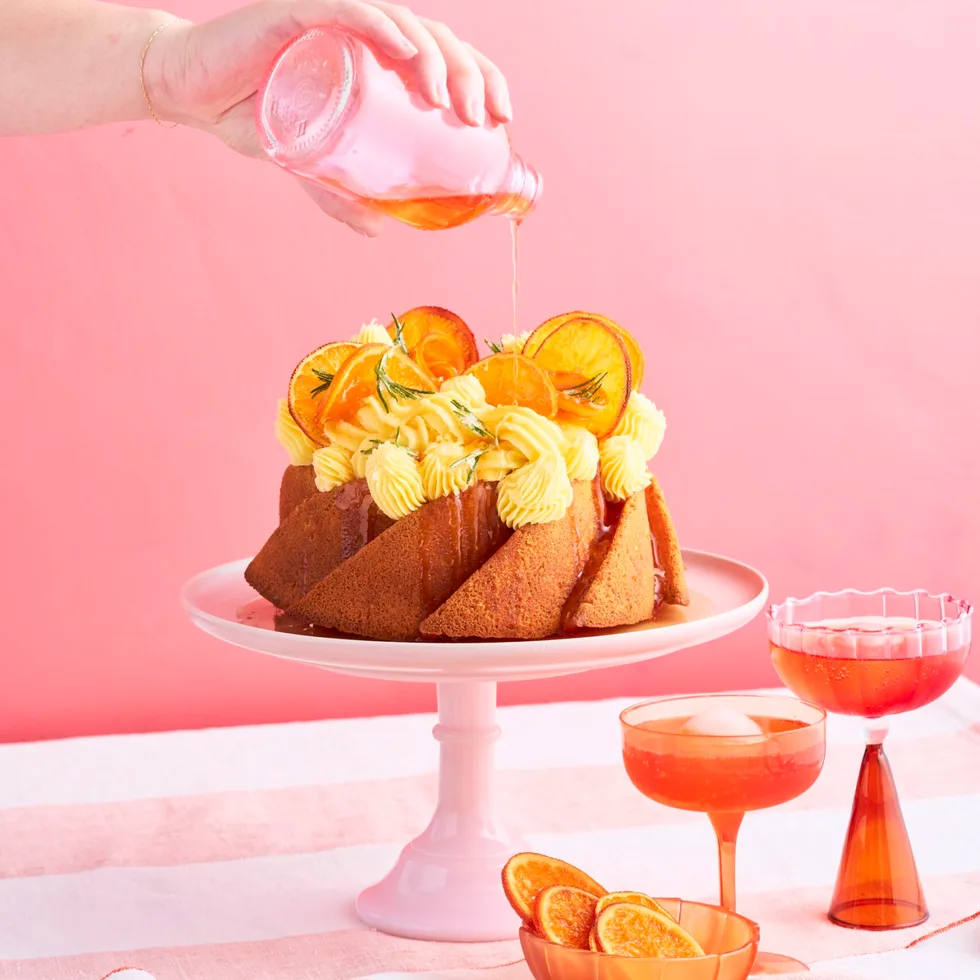 A round naked budt cake with cream ontop and garnished with orange slices sitting ontop of a light pink cake stand. A hand is hovering from above the cake drizzling a aperol syrup ontop of the cake.