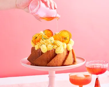 A round naked budt cake with cream ontop and garnished with orange slices sitting ontop of a light pink cake stand. A hand is hovering from above the cake drizzling a aperol syrup ontop of the cake.