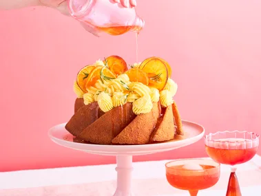 A round naked budt cake with cream ontop and garnished with orange slices sitting ontop of a light pink cake stand. A hand is hovering from above the cake drizzling a aperol syrup ontop of the cake.