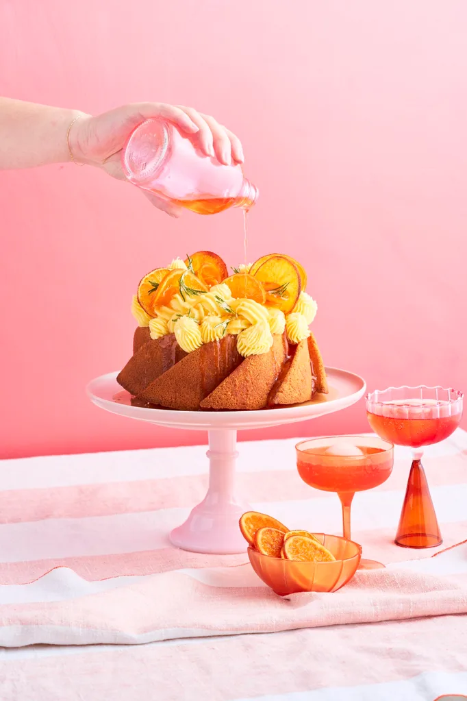 A round naked budt cake with cream ontop and garnished with orange slices sitting ontop of a light pink cake stand. A hand is hovering from above the cake drizzling a aperol syrup ontop of the cake.