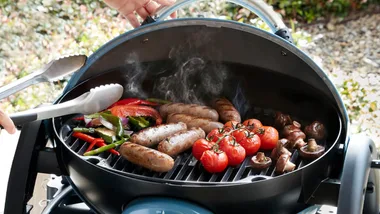 An image of a portable bbq with an open hood cooking sausages, tomatoes and other vegetables on the grill with tongs.