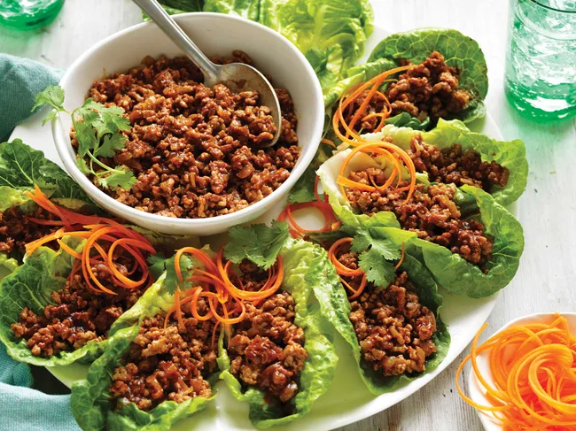 Lettuce wraps filled with seasoned ground meat, garnished with carrot spirals and cilantro, with a bowl of filling.
