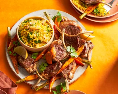 lamb cutlets thrown on baby carrots with small bowl on plate with rice in it