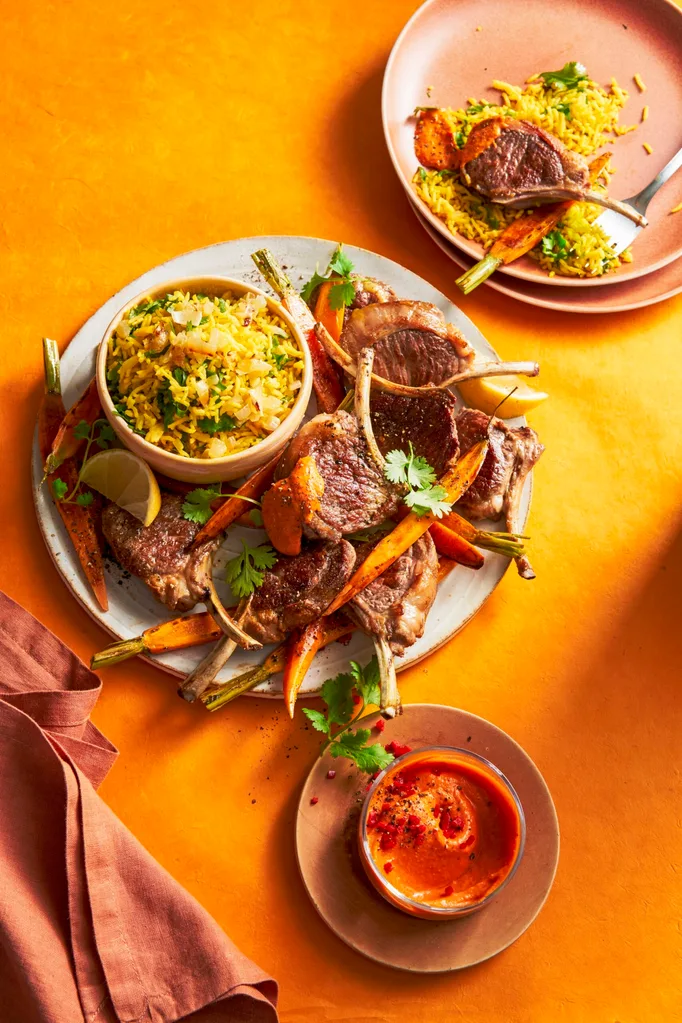lamb cutlets thrown on baby carrots with small bowl on plate with rice in it