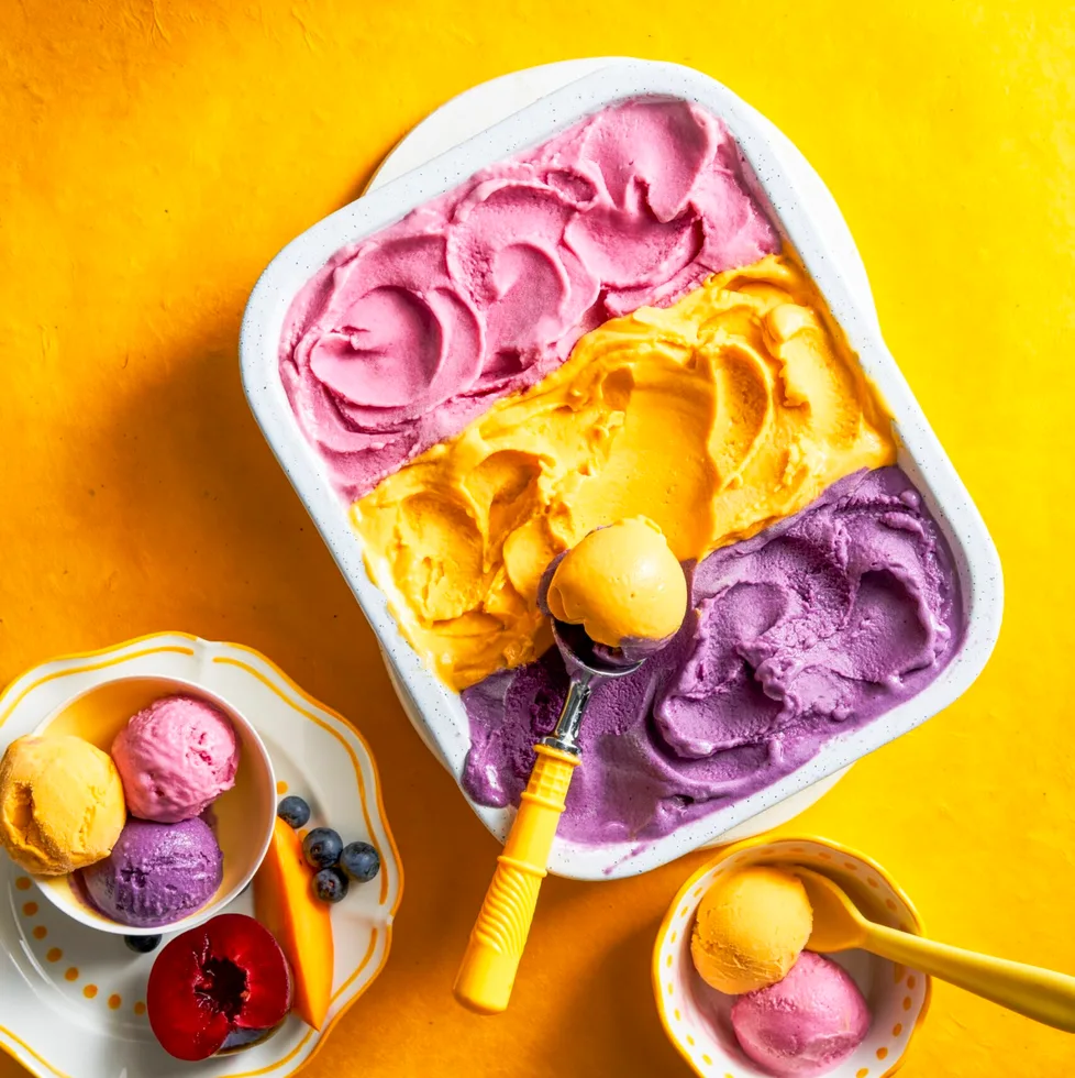 three flavours of gelato with purple, yellow and pink in one container with spoon scooping on top