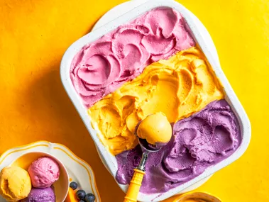 three flavours of gelato with purple, yellow and pink in one container with spoon scooping on top