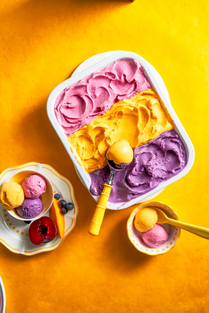 three flavours of gelato with purple, yellow and pink in one container with spoon scooping on top
