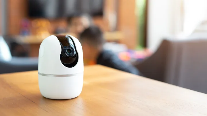 Small white security camera on a wooden table, with blurred-out children playing in their living room in the background.