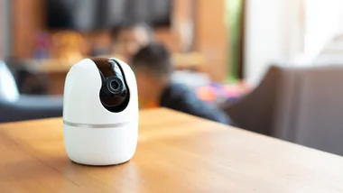 Small white security camera on a wooden table, with blurred-out children playing in their living room in the background.
