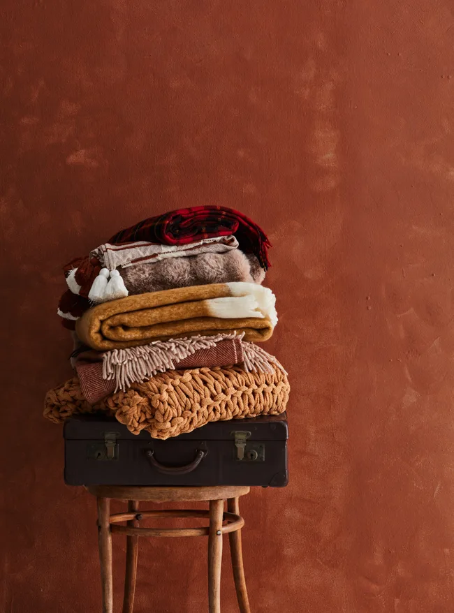 throw blankets folded up messily and placed in pile on wooden stool. background is deep terracotta red backdrop
