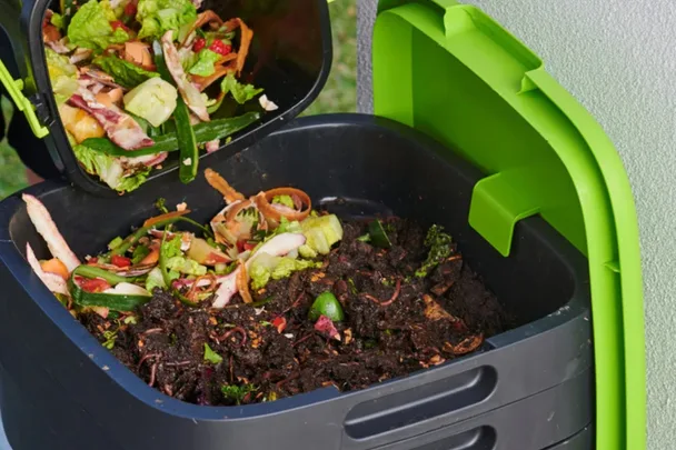 compost bin with green lid open and fresh vegetable scraps being poured into fresh soil