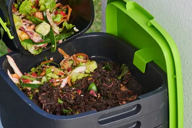 compost bin with green lid open and fresh vegetable scraps being poured into fresh soil