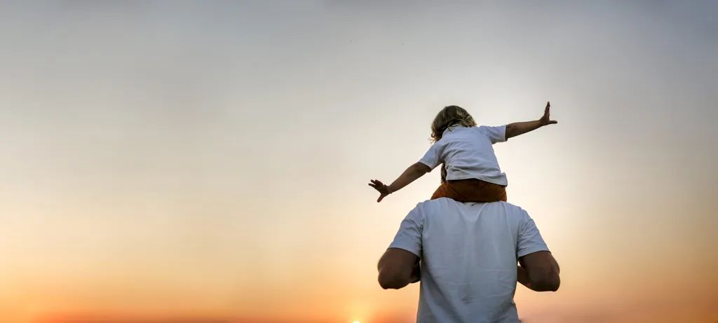 child on the shoulders of father not facing the camera with arms out like an airplane, sunset in the background