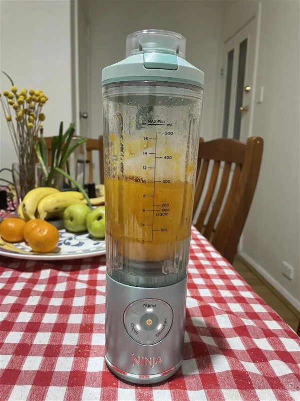 ninja portable blender with mandarin and sugar inside blending and fruit bowl behind. on top of red gingham tablecloth in dining room