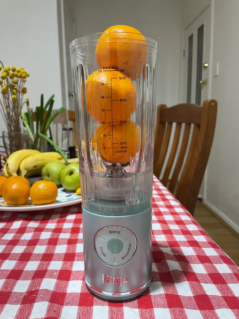 ninja portable blender with three stacked mandarins inside and fruit bowl behind. on top of red gingham tablecloth in dining room
