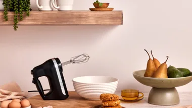 Kitchen items on a bench with a hand mixer in front, set against a pink background, available with KitchenAid discount codes