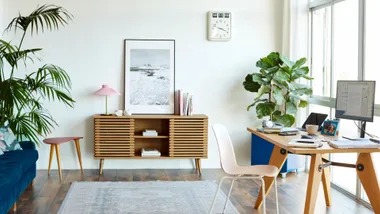 Modern home office with a wooden desk, desktop monitor and indoor plants, featuring mid-century furniture and a large window letting in natural light.