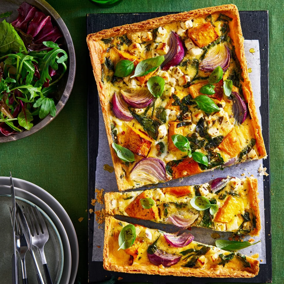roasted pumpkin quiche cut in half crookedly sitting on baking tray with leafy herbs in bowl in corner