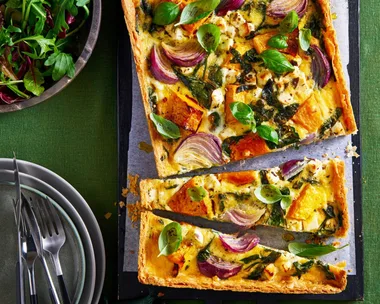 roasted pumpkin quiche cut in half crookedly sitting on baking tray with leafy herbs in bowl in corner