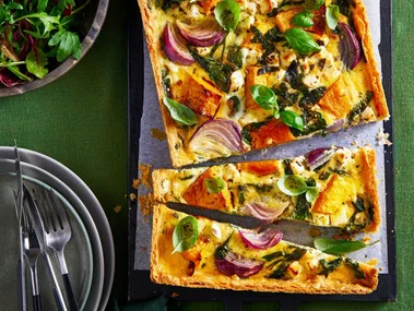 roasted pumpkin quiche cut in half crookedly sitting on baking tray with leafy herbs in bowl in corner