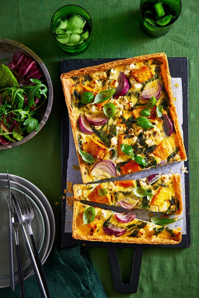 roasted pumpkin quiche cut in half crookedly sitting on baking tray with leafy herbs in bowl in corner
