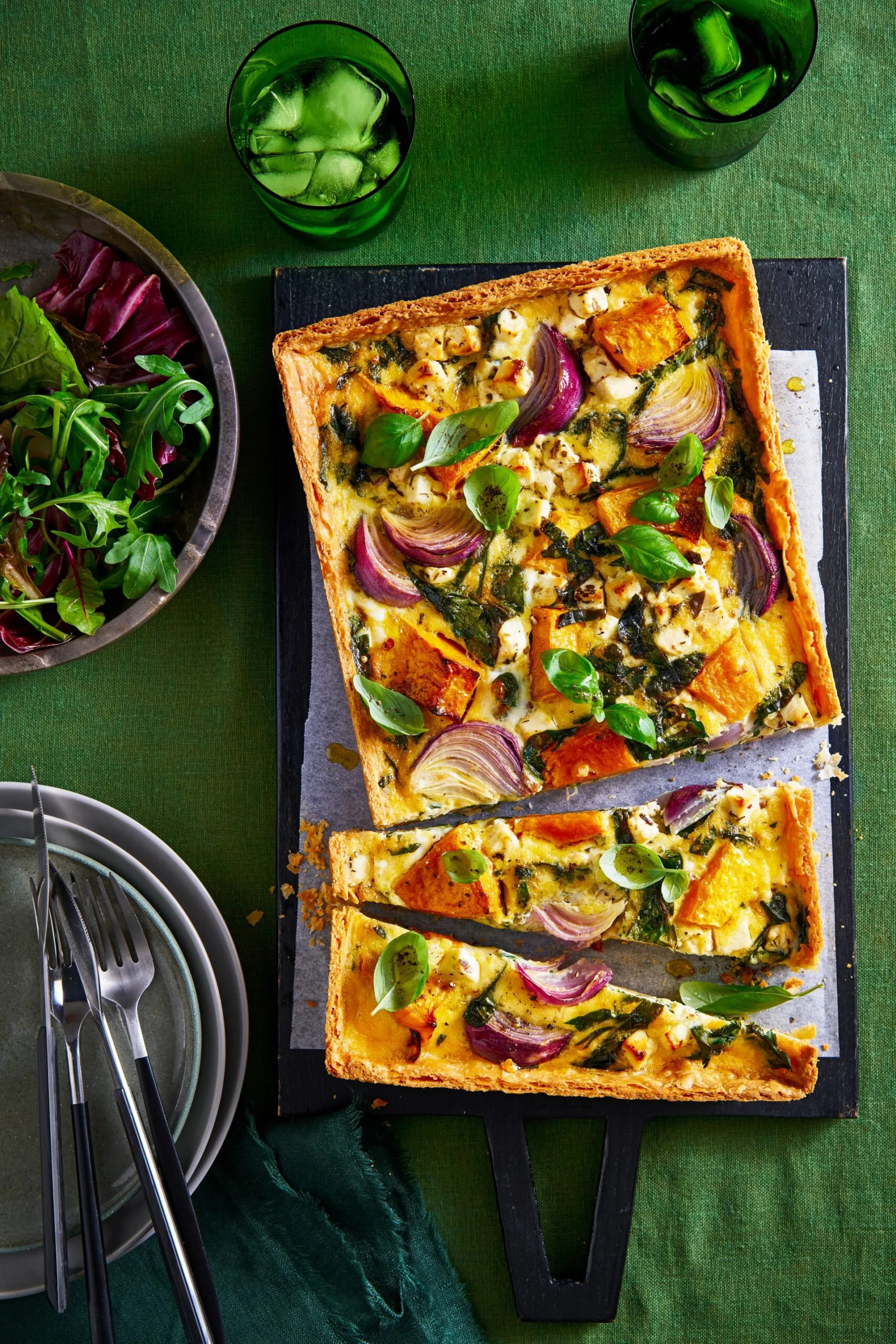 roasted pumpkin quiche cut in half crookedly sitting on baking tray with leafy herbs in bowl in corner