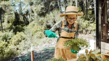 An older woman with glasses and a straw hat smiles while gardening. She is wearing a plaid shirt and tan overalls with green gloves, and is holding a small potted plant in one hand and a gardening trowel in the other. She is standing outdoors on a sunny day.