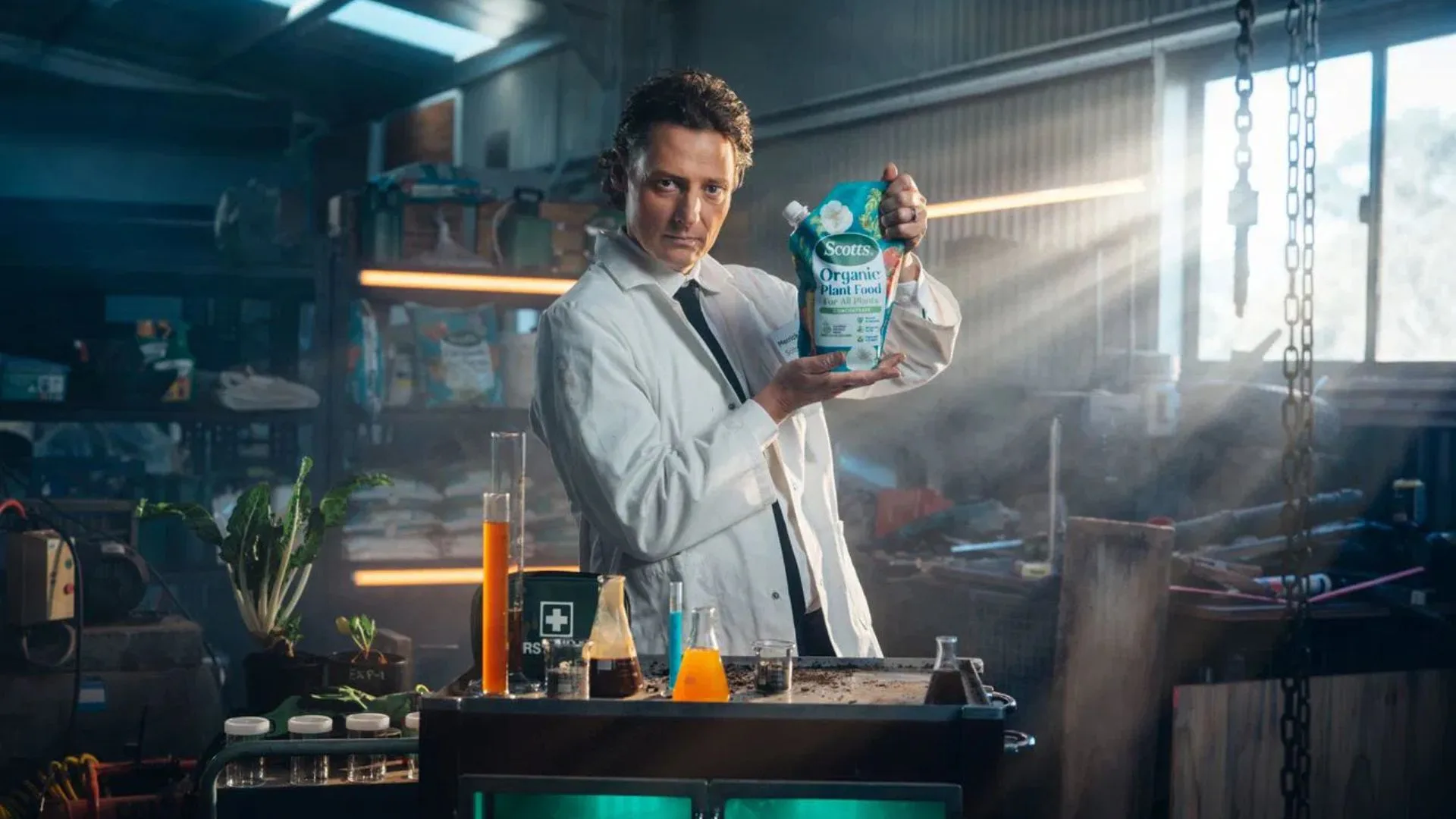 Comedian Merrick Watts, dressed as a 'Soilmelier' in a lab coat, holds up a bag of Scotts Organic Plant Food in a cluttered shed laboratory.