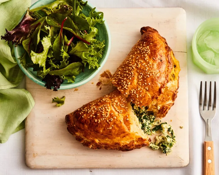 spinach and cheese hand pie cut in half with filling spilling out with fork and bowl of salad on the side