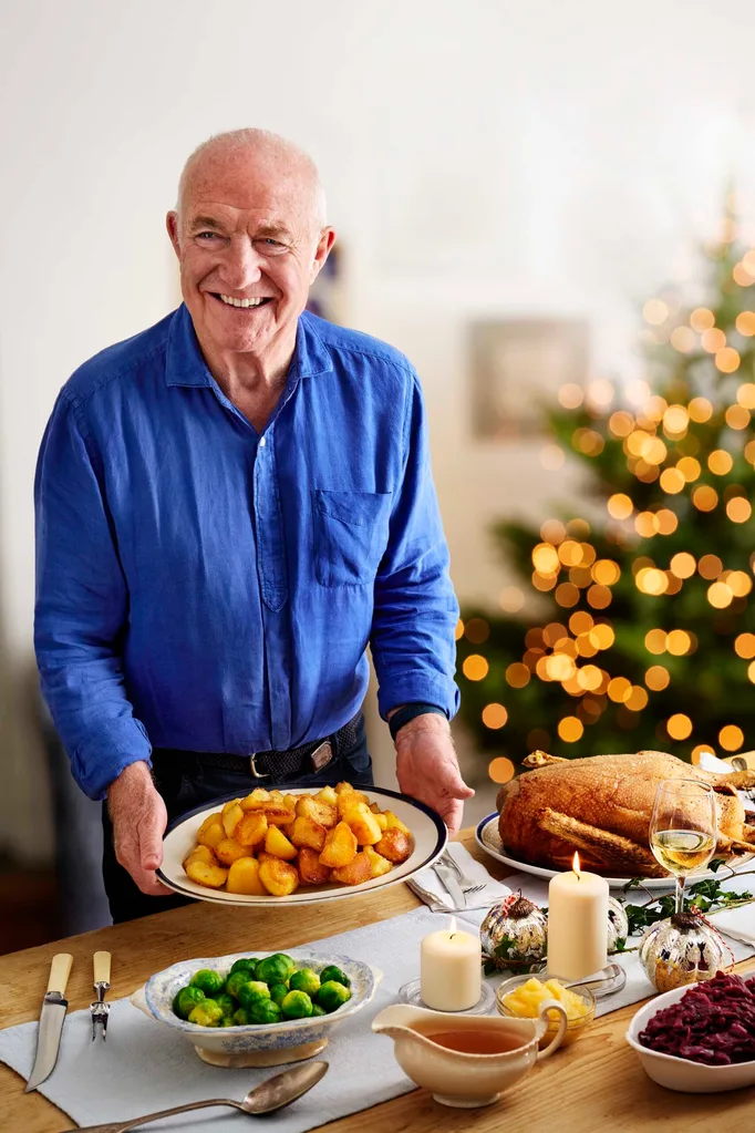 Chef Rick Stein wearing a blue shirt and placing a dish on a table laid out for Christmas