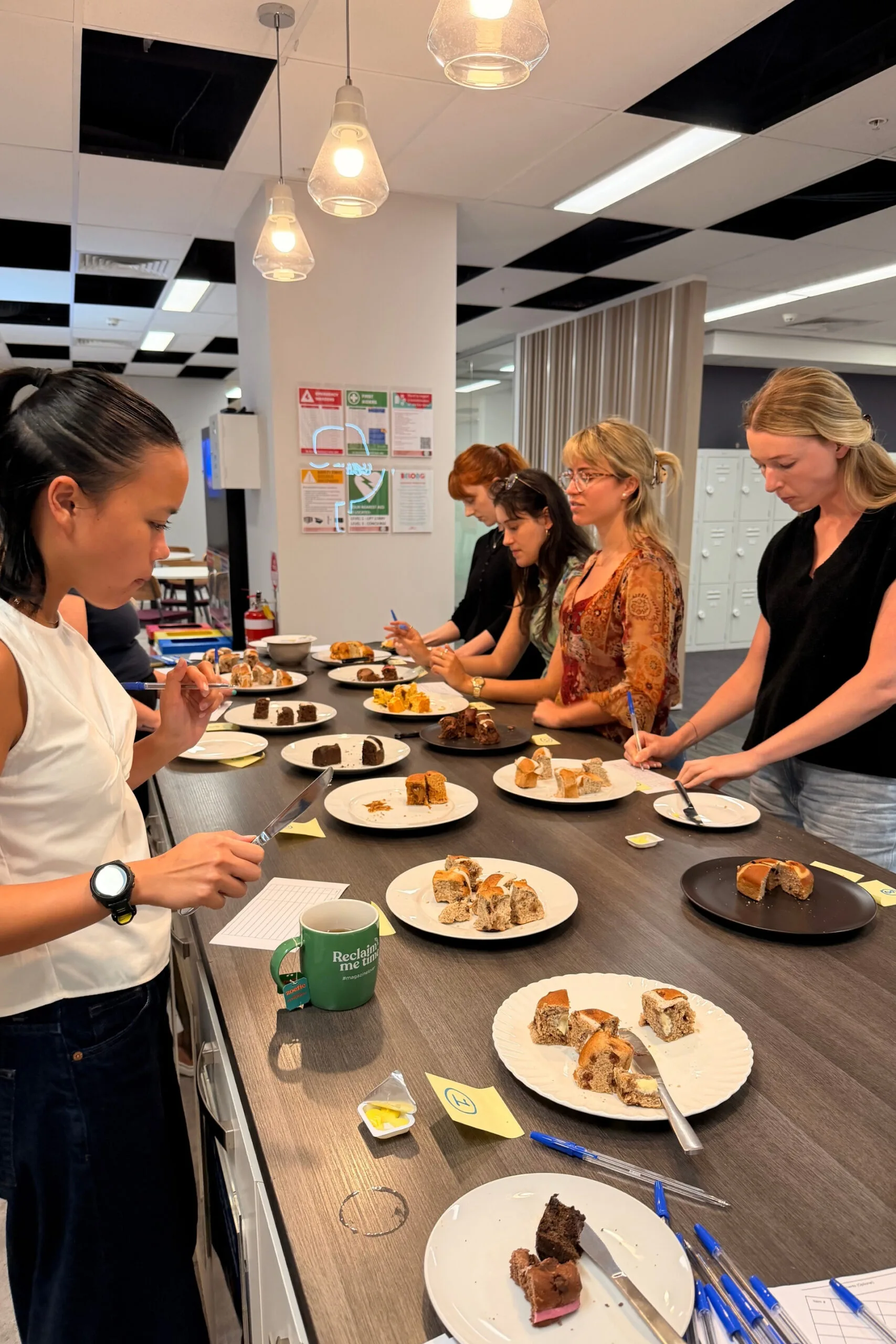 four women on one side of bench taste testing supermarket hot cross buns in office kitchen and one woman on left side with cup of tea