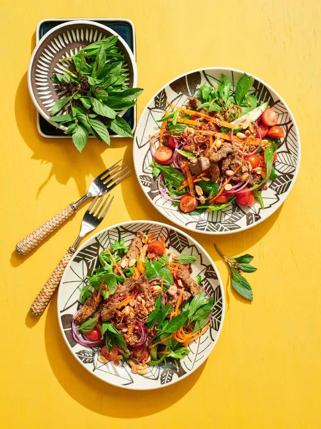 thai beef salad with beef and coriander on top of leafy green salad with mint and coriander on separate plate on the side
