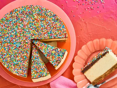 no bake freckly cheesecake on pink plate with several slices cut into it. one slice appears on the right on a peach fluted plate, background is pink to orange ombre