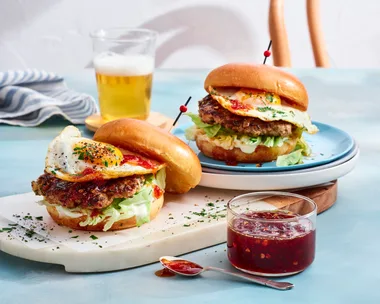 two breakfast burgers with pork smash pattie and eggs on them. chilli jam in clear jar to the front and glass of orange juice at the back