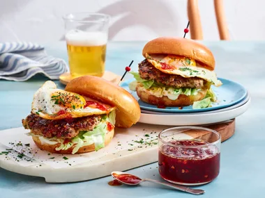 two breakfast burgers with pork smash pattie and eggs on them. chilli jam in clear jar to the front and glass of orange juice at the back