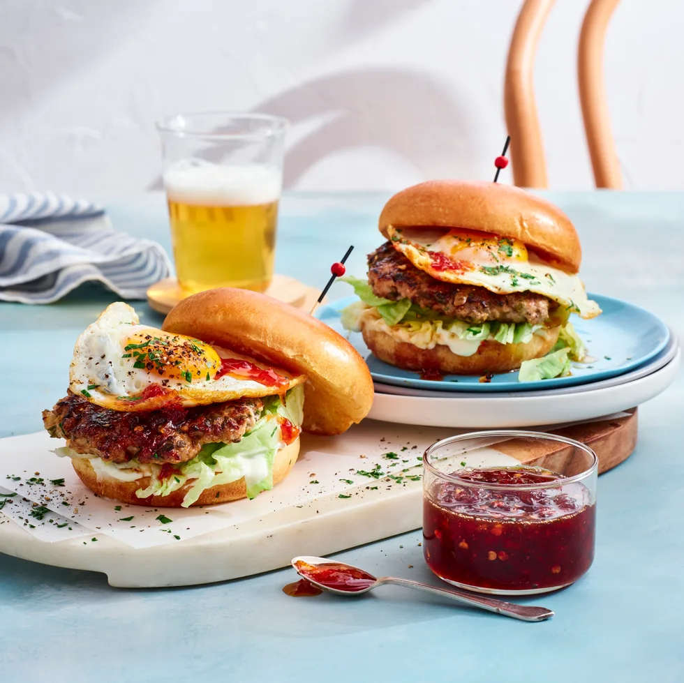 two breakfast burgers with pork smash pattie and eggs on them. chilli jam in clear jar to the front and glass of orange juice at the back