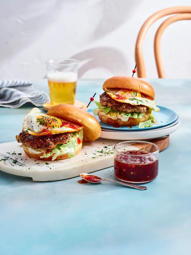 two breakfast burgers with pork smash pattie and eggs on them. chilli jam in clear jar to the front and glass of orange juice at the back