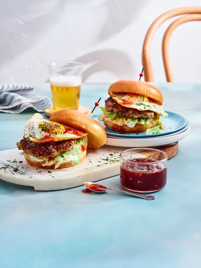 two breakfast burgers with pork smash pattie and eggs on them. chilli jam in clear jar to the front and glass of orange juice at the back