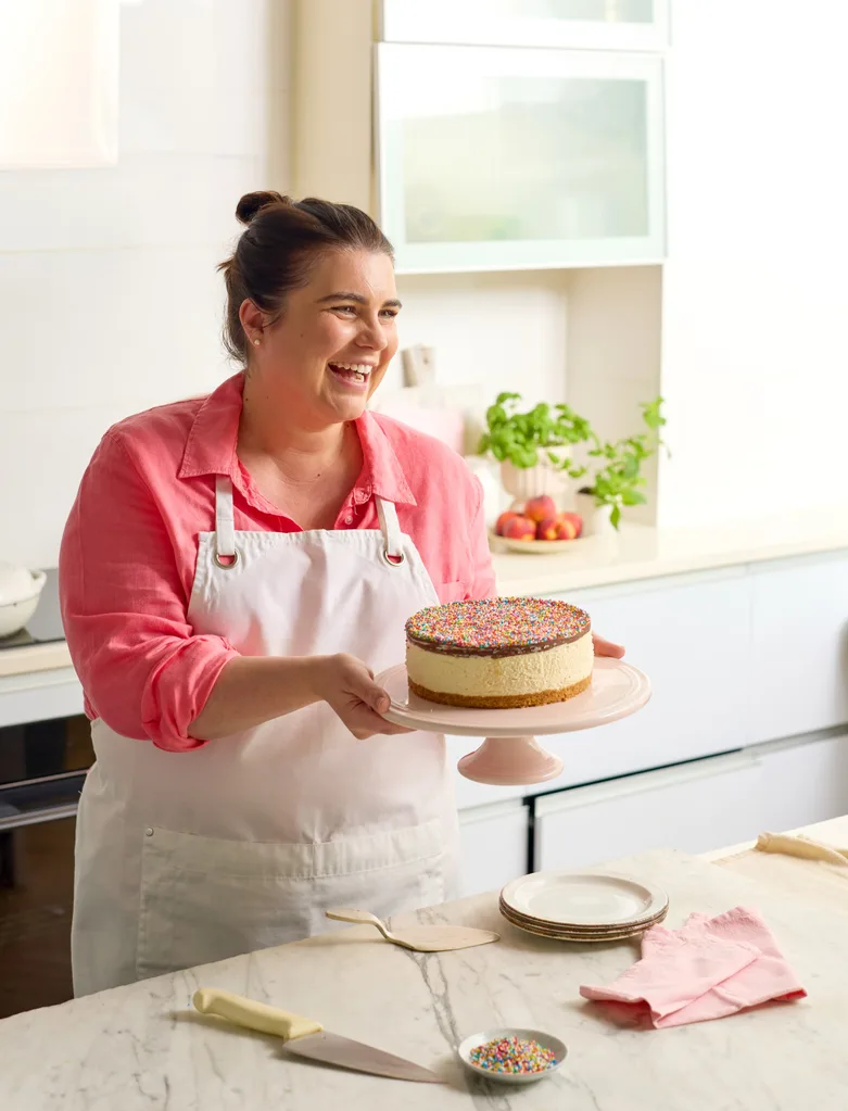 sarah murphy holding up freckle no bake cheesecake on cake stand wearing a pink shirt and white apron
