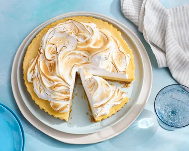 yuzu meringue pie with one slice cut out and browned meringue topping. On grey plate with dappled blue tablecloth underneath.