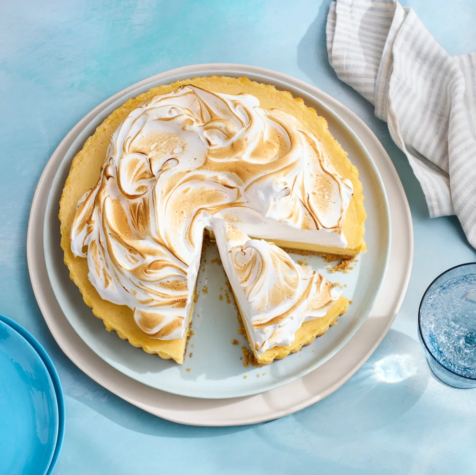 yuzu meringue pie with one slice cut out and browned meringue topping. On grey plate with dappled blue tablecloth underneath.