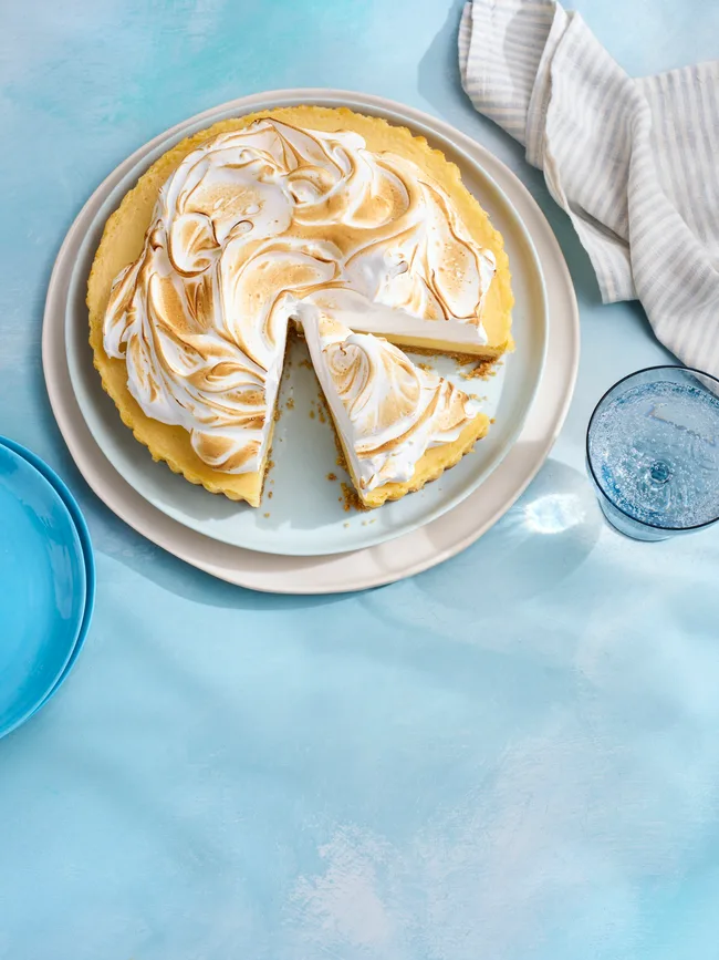 yuzu meringue pie with one slice cut out and browned meringue topping. On grey plate with dappled blue tablecloth underneath.