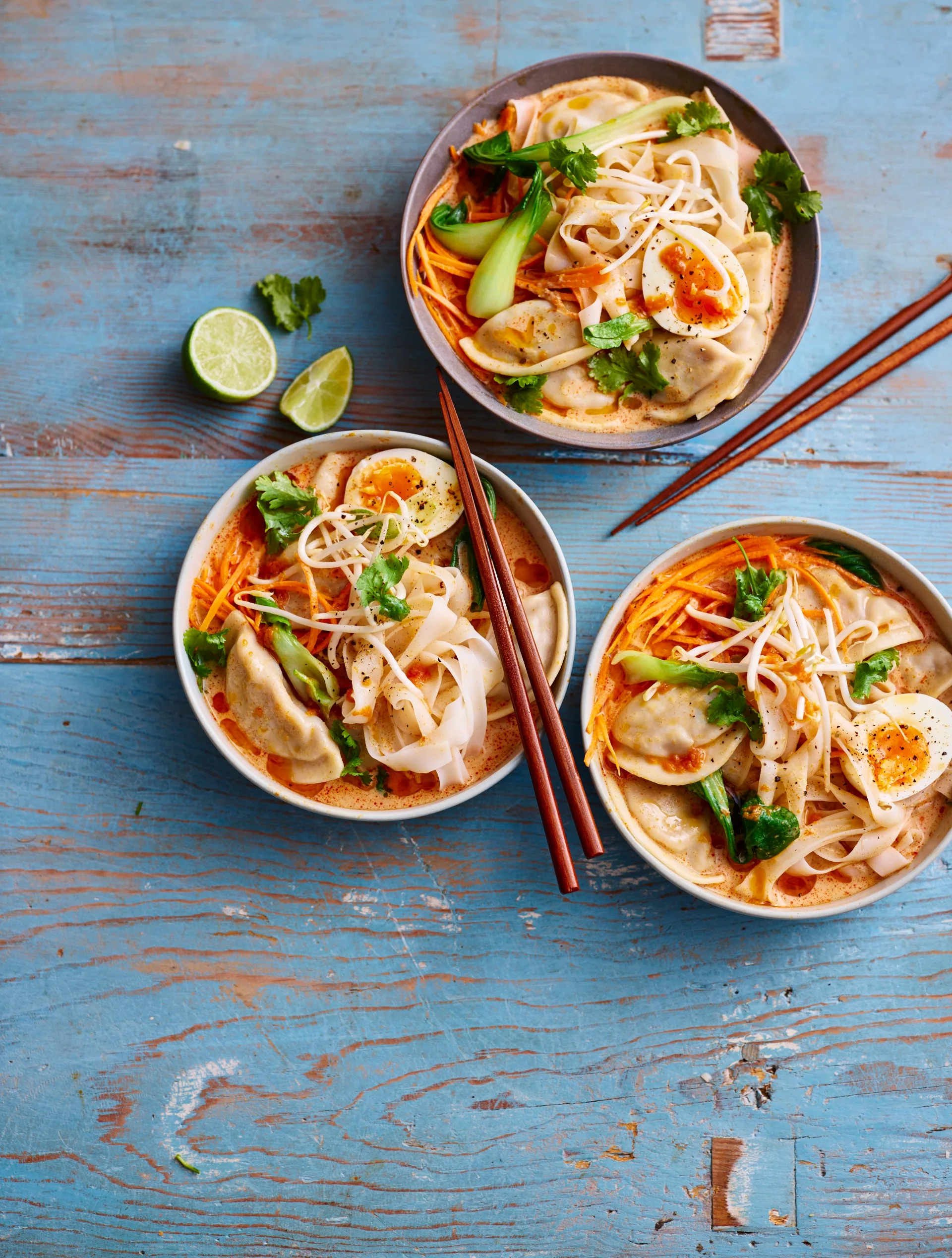 dumpling laksa in three bowls with lime wedges on the side and coriander on top