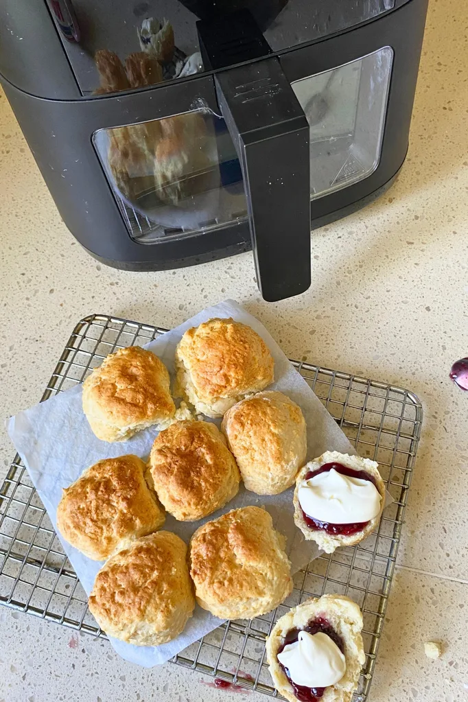 Air fryer lemonade scones on a cooling rack next to an air fryer. One scone has been cut open and has jam and cream on it.